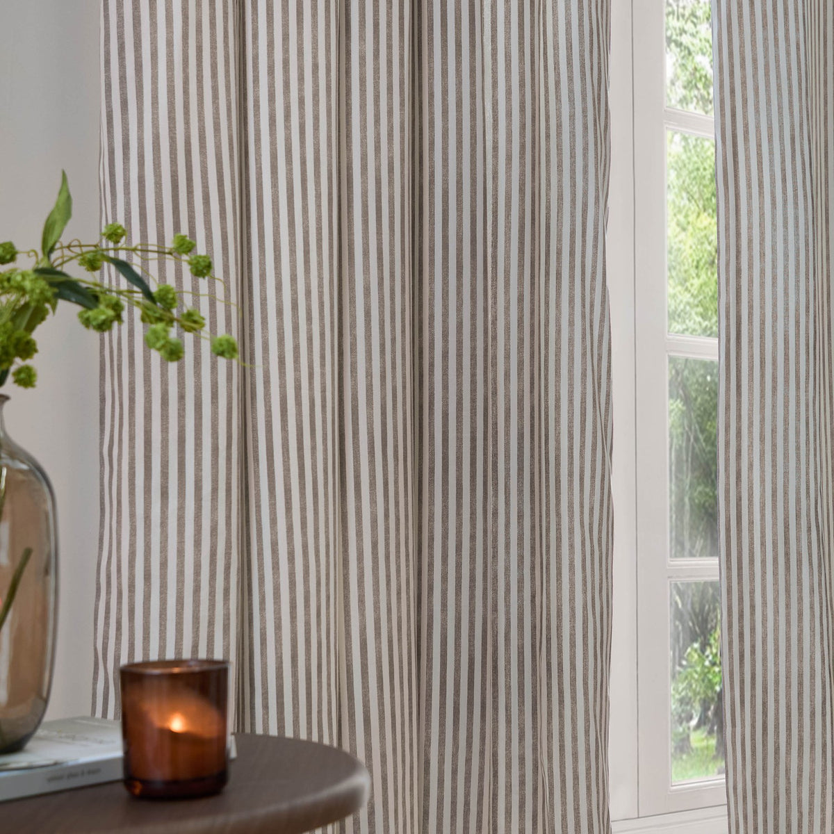 Hebden Striped Cotton Pencil Pleat Curtains in mole-brown stripe on cream, hung floor-length beside a window, styled with a glass vase and candle on a side table.
