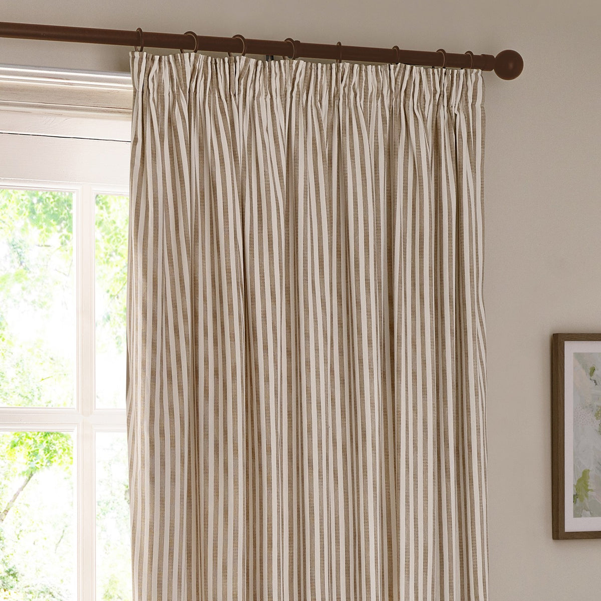 Hebden pencil pleat curtains in natural cotton stripe, hung on a dark wood pole with rings, shown at a window in a light-toned room.