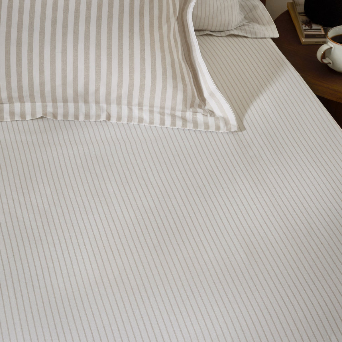 Hebden Striped Cotton Fitted Sheet in Natural on a bedroom mattress, showing narrow sandy taupe stripes on a cream cotton ground, with matching striped pillowcases behind.