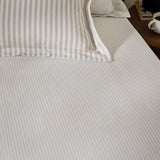 Hebden Striped Cotton Fitted Sheet in Natural on a bedroom mattress, showing narrow sandy taupe stripes on a cream cotton ground, with matching striped pillowcases behind.