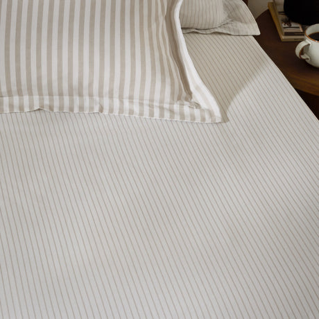 Hebden Striped Cotton Fitted Sheet in Natural on a bedroom mattress, showing narrow sandy taupe stripes on a cream cotton ground, with matching striped pillowcases behind.