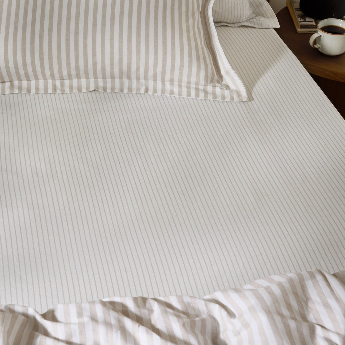 Hebden Striped Cotton Fitted Sheet in natural on a bedroom mattress, showing narrow taupe stripes on cream cotton with matching pillowcase and duvet.