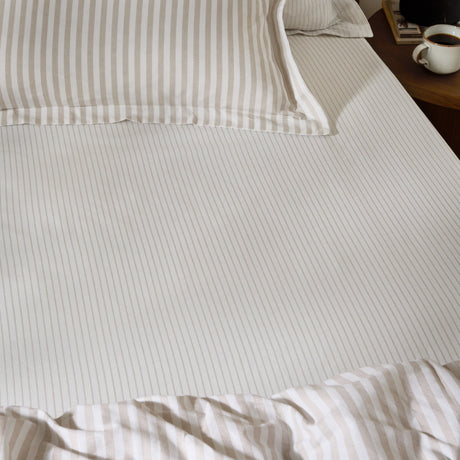 Hebden Striped Cotton Fitted Sheet in natural on a bedroom mattress, showing narrow taupe stripes on cream cotton with matching pillowcase and duvet.
