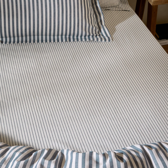 Hebden striped cotton fitted sheet in navy, shown fitted on a bed with matching striped pillowcases and duvet, fine navy stripes on an ivory ground.