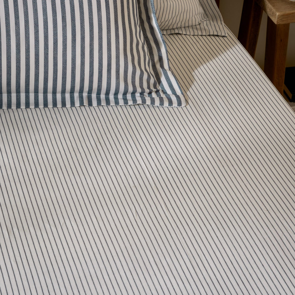 Hebden fitted sheet in navy on a bed, close-set narrow stripes on pale ivory cotton, with matching striped pillowcases visible at the headboard.