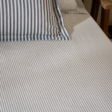 Hebden fitted sheet in navy on a bed, close-set narrow stripes on pale ivory cotton, with matching striped pillowcases visible at the headboard.
