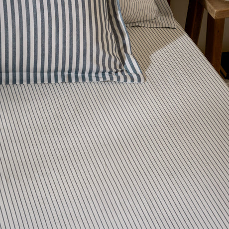 Hebden fitted sheet in navy on a bed, close-set narrow stripes on pale ivory cotton, with matching striped pillowcases visible at the headboard.
