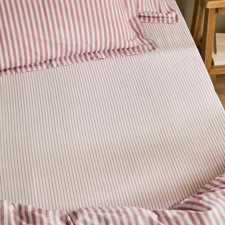 Hebden fitted sheet in 100% cotton with a fine rosewood pink and cream stripe, shown fitted on a bedroom mattress alongside matching pillowcases.