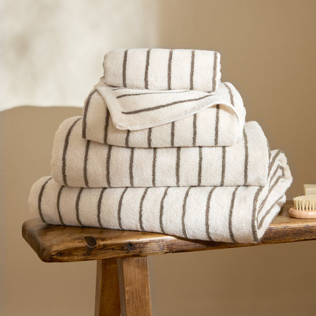 Hebden Striped Cotton Towels in Mole, stacked in varying sizes with cream and brown stripes, shown on a wooden bench in a bathroom setting.