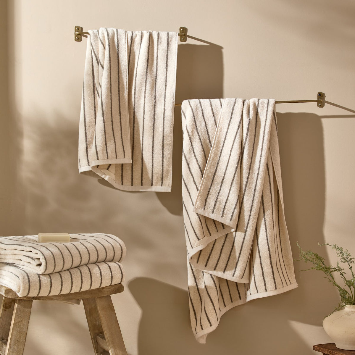Hebden Striped Cotton Towels in Mole hang from wall-mounted brass rails, with folded matching towels stacked on a wooden stool, in a warm neutral bathroom setting.
