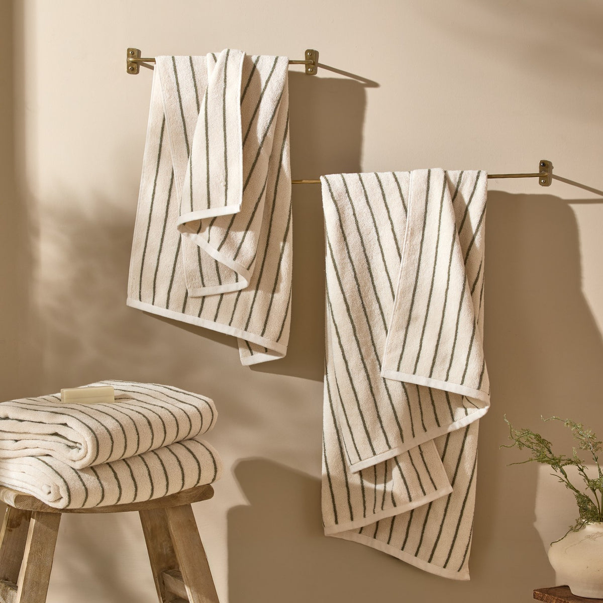 Hebden Striped Cotton Towels in olive on cream, shown hanging from brass wall rails and folded on a wooden stool in a bathroom setting.