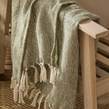 Hedon Striped Polyester Throw in moss green, woven with a subtle two-tone stripe and knotted fringe, draped over the wooden arm of a chair.