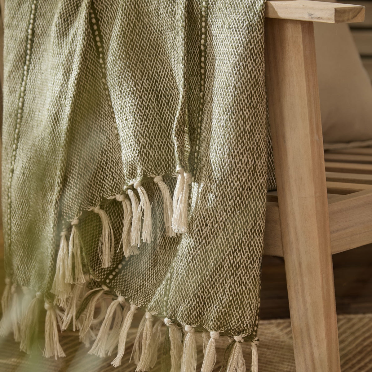 Hedon Striped Polyester Throw in moss green draped over a wooden chair arm, showing a two-tone woven texture with knotted fringe tassels.