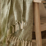 Hedon Striped Polyester Throw in moss green draped over a wooden chair arm, showing a two-tone woven texture with knotted fringe tassels.