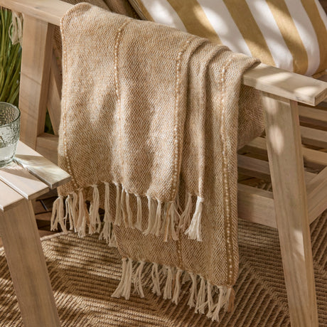 Hedon Striped Polyester Throw in natural sand tone with woven texture, draped over a wooden outdoor chair with knotted fringe edges visible.