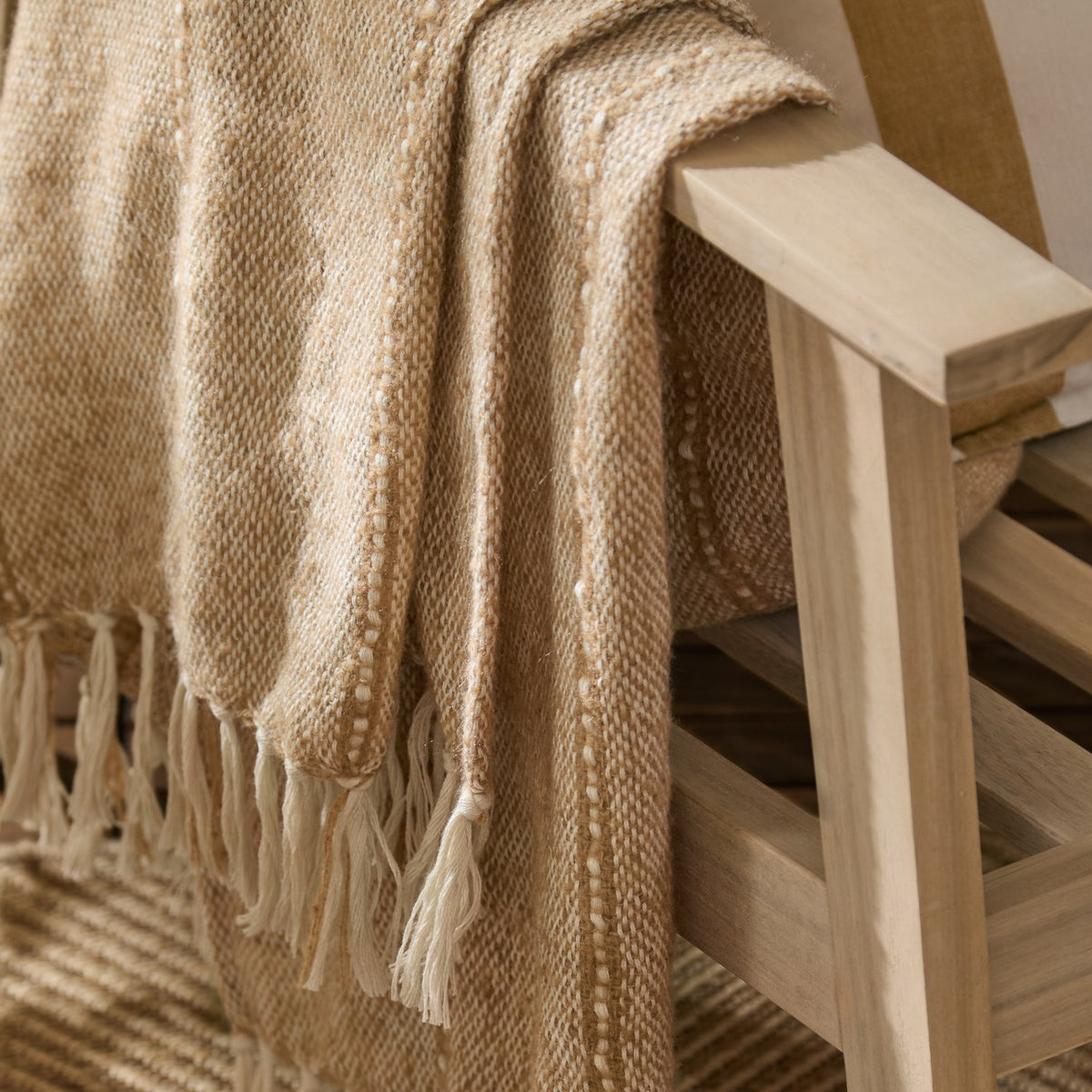 Hedon Striped Polyester Throw in Natural draped over a wooden chair arm, showing woven texture with subtle stripes and knotted fringe detail.