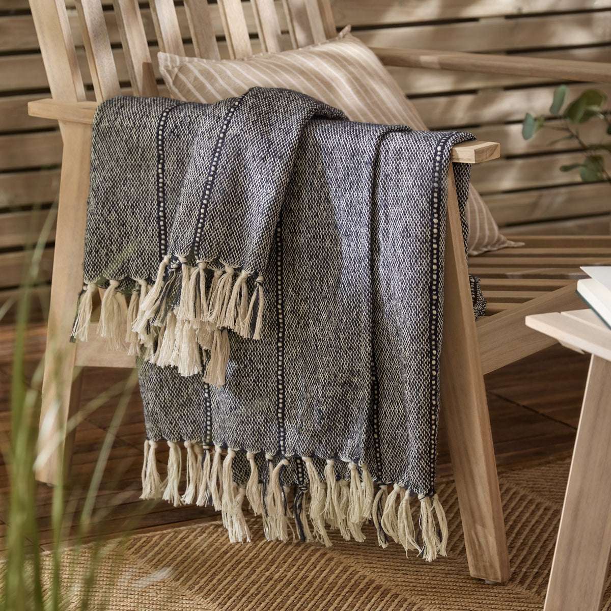 Hedon Striped Polyester Throw in navy and cream marl with knotted fringe, draped over a wooden armchair beside a striped cushion in an outdoor living space.