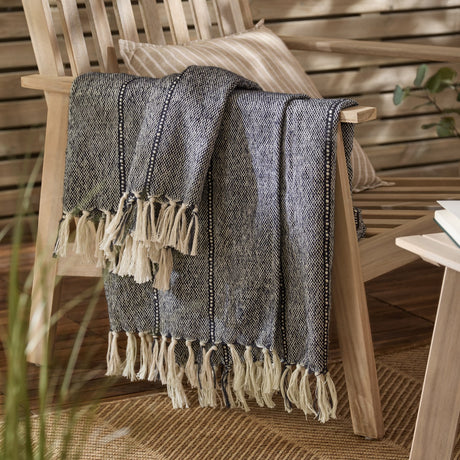 Hedon Striped Polyester Throw in navy and cream marl with knotted fringe, draped over a wooden armchair beside a striped cushion in an outdoor living space.