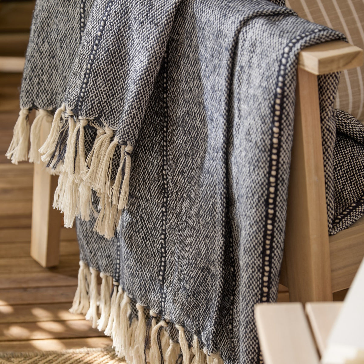 Hedon Striped Polyester Throw in navy and cream marl with knotted fringe, draped over a wooden armchair on an outdoor deck.