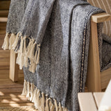 Hedon Striped Polyester Throw in navy and cream marl with knotted fringe, draped over a wooden armchair on an outdoor deck.