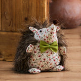 Henrietta Polyester Door Stop shaped as a hedgehog with faux fur spikes, floral cream fabric face and belly, olive-green bow, and a brown pom-pom nose, shown on a wooden floor.