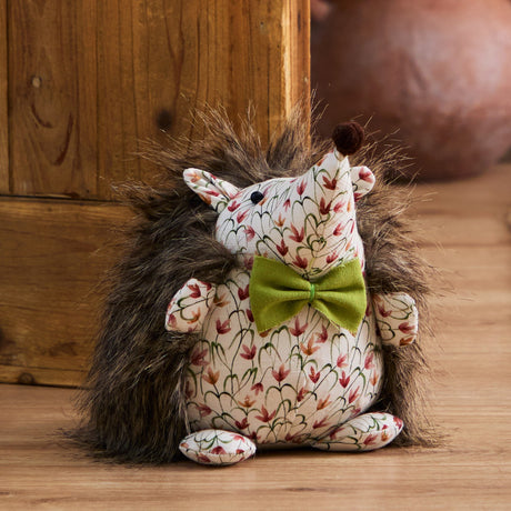 Henrietta Polyester Door Stop shaped as a hedgehog with faux fur spikes, floral cream fabric face and belly, olive-green bow, and a brown pom-pom nose, shown on a wooden floor.