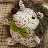 Henrietta hedgehog-shaped door stop with cream floral fabric body, shaggy brown faux fur back, black bead eyes, brown pom-pom nose, and an olive-green bow at the neck.