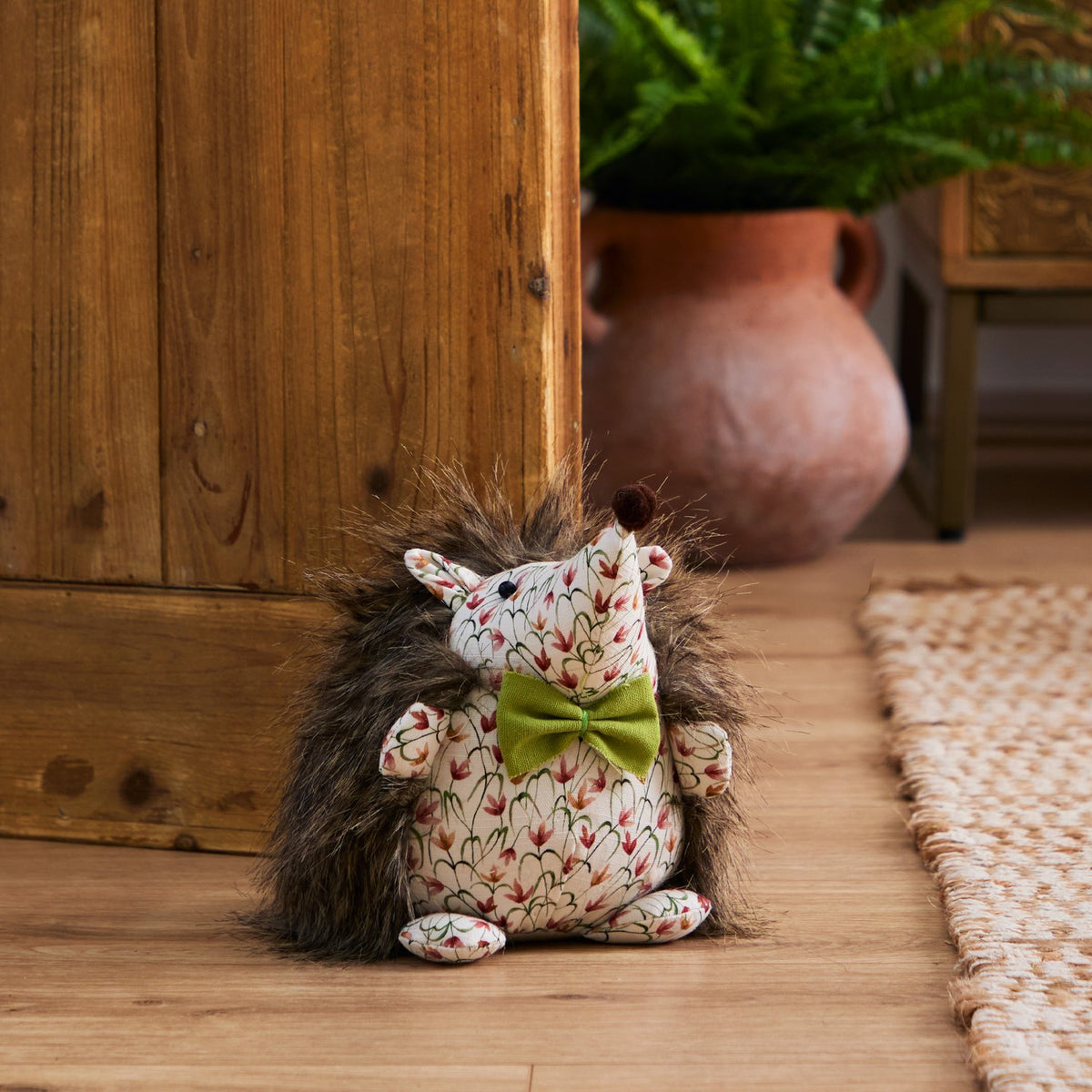 Henrietta Polyester Door Stop shaped like a hedgehog with faux fur back, cream floral fabric body, olive-green bow, and brown pom-pom nose, set on a wooden floor by a door.