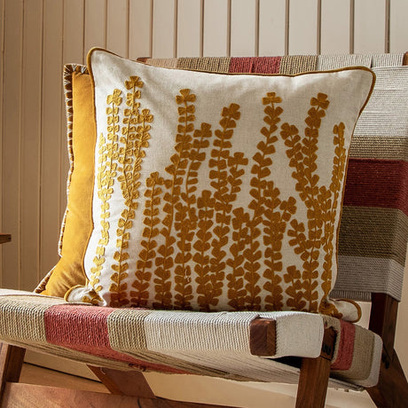 Herb Velvet Linen Cushion in mustard, square with embroidered botanical stem design on a cotton-linen front, shown on a striped chair beside a plain mustard velvet cushion.