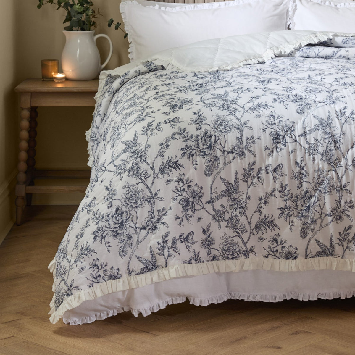 Hermione Linen Cotton Bedspread in Ivory Blue on a bed, featuring an inky blue Toile de Jouy floral print on cream with a ruffled edge, shown in a bedroom setting.