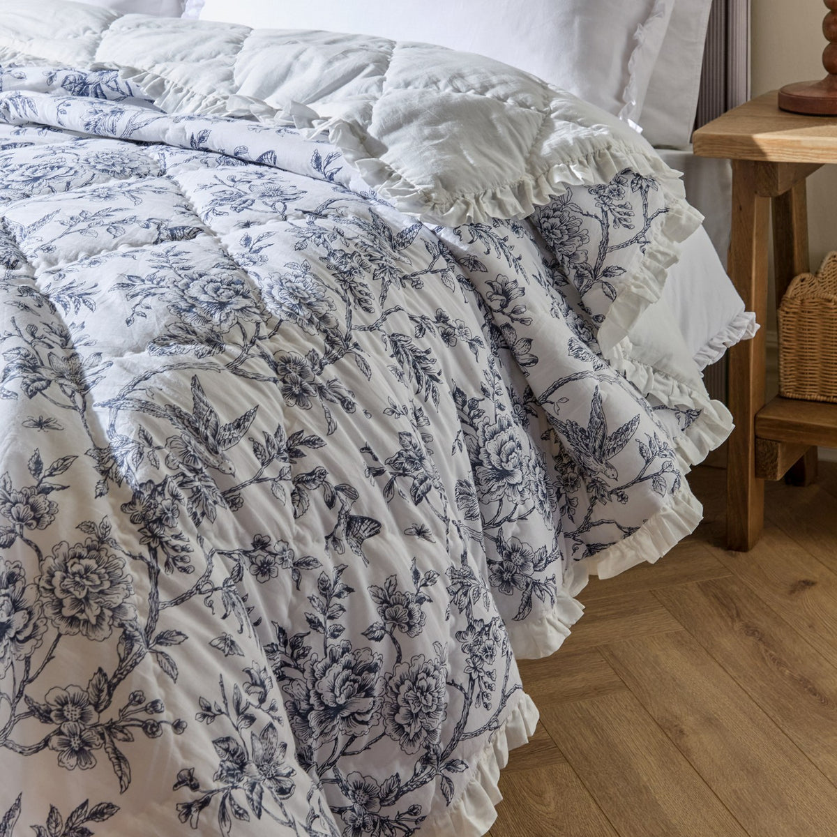 Hermione linen cotton bedspread in ivory blue, quilted diamond design with blue floral toile pattern and ruffled edge, shown on a bed beside a wooden nightstand.