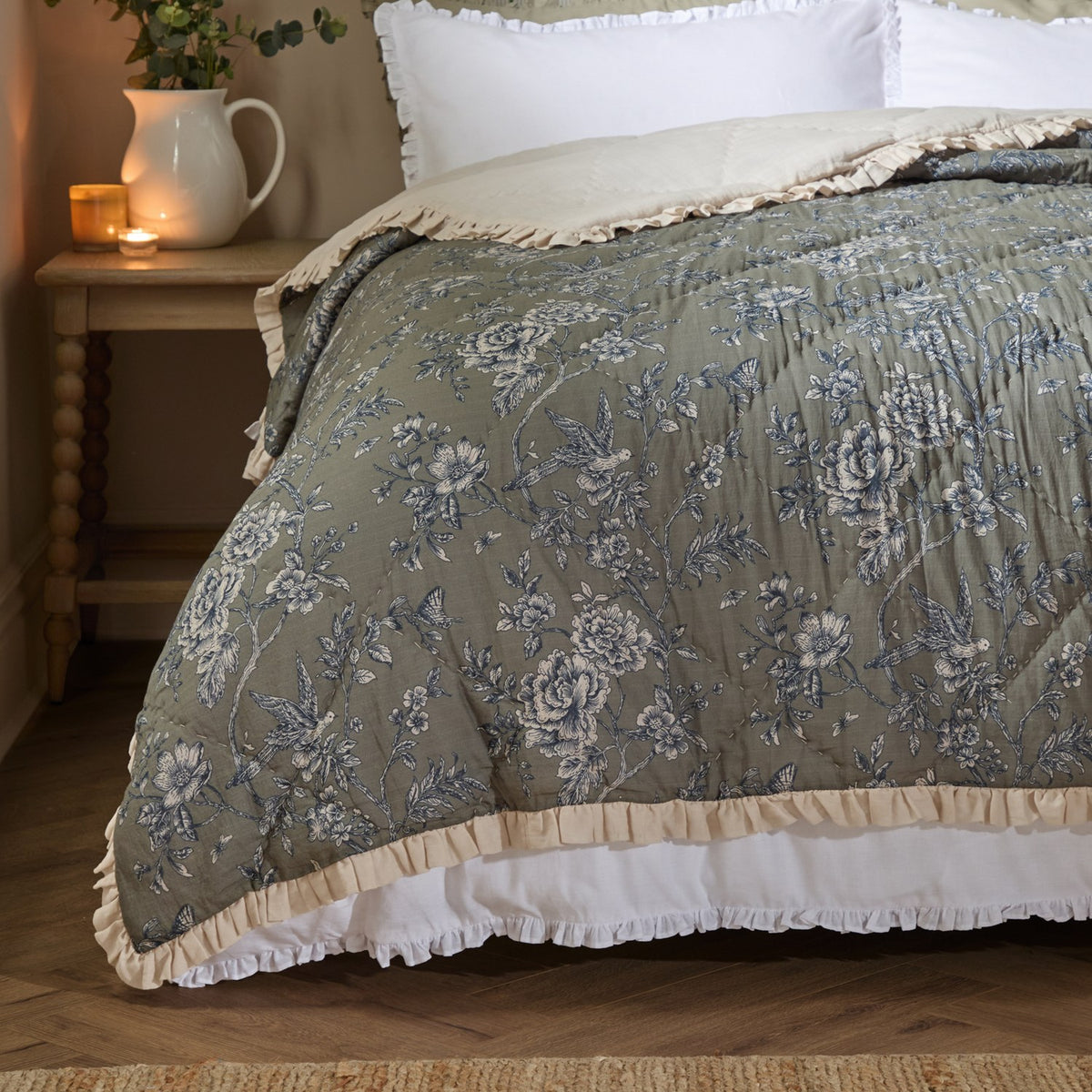 Quilted Hermione linen cotton bedspread in moss with a blue and cream Toile de Jouy floral, bird and butterfly print and cream ruffled frill, dressing a bed beside a wooden nightstand.