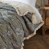Hermione Linen Cotton Bedspread in Moss, quilted with a blue and cream floral toile print, cream ruffled frill edging, shown folded on a bed beside a wooden nightstand.