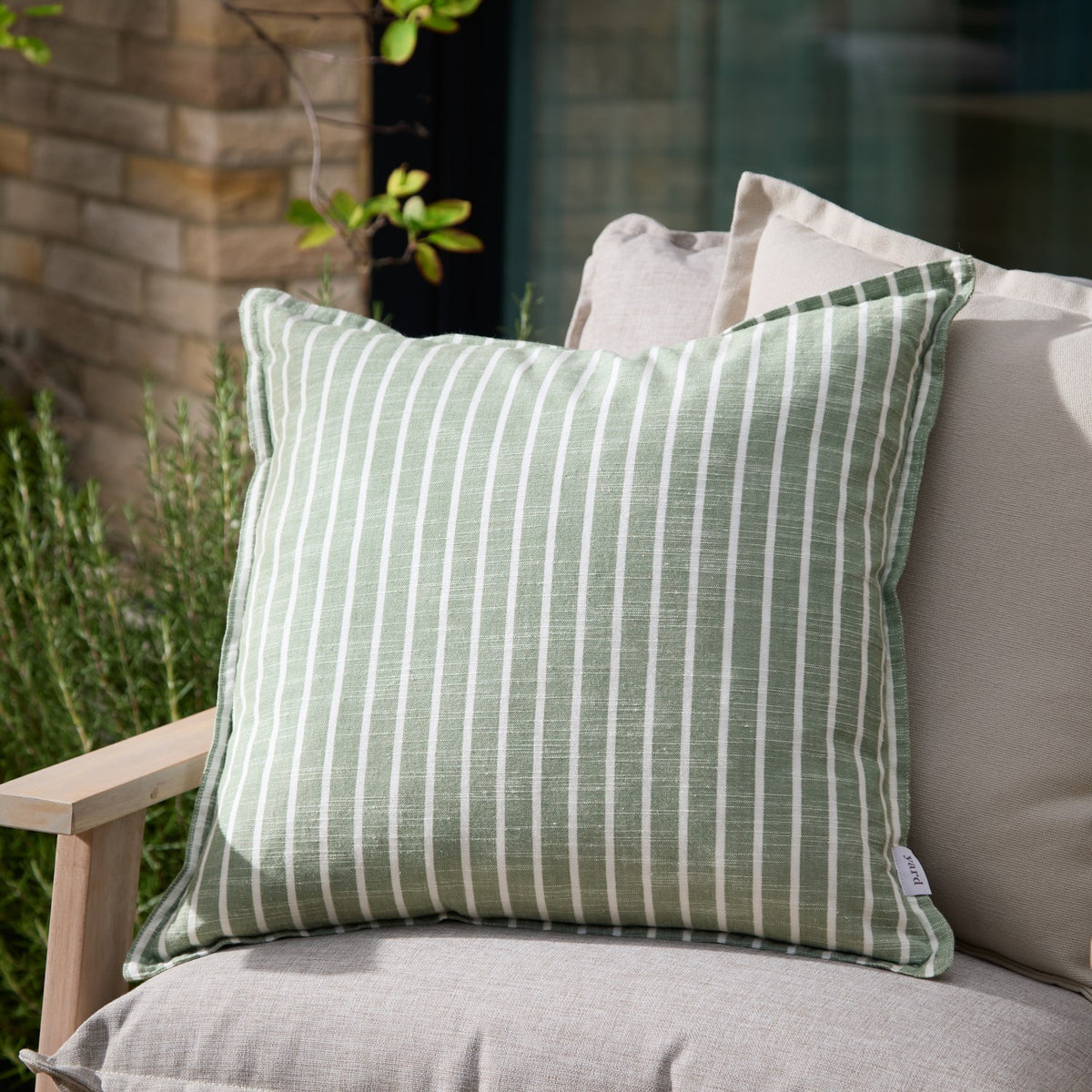 Hessle Stripe Yarn-Dyed Cotton Cushion in eucalyptus green with white pinstripes, shown on an outdoor sofa alongside neutral cushions in a garden setting.