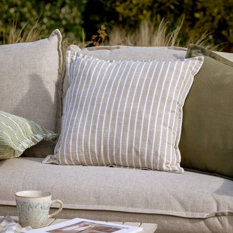 Hessle Stripe Yarn-Dyed Cotton Cushion in natural with white vertical stripes, shown on a beige outdoor sofa alongside olive green cushions.