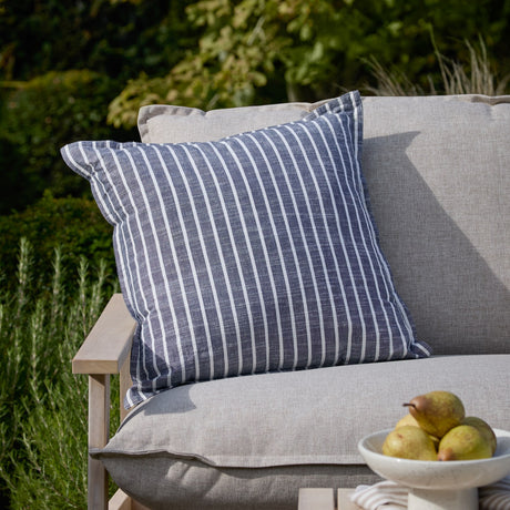 Hessle Stripe Yarn-Dyed Cotton Cushion in navy with white stripes and an oxford edge, shown on a grey outdoor sofa in a garden setting.