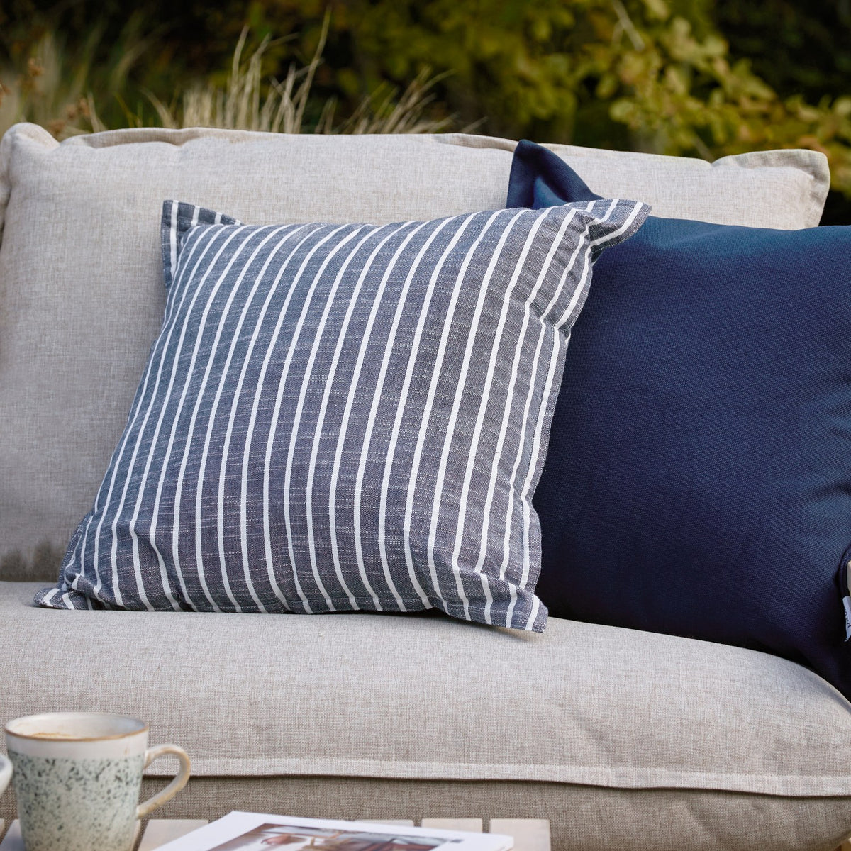 Hessle Stripe Yarn-Dyed Cotton Cushion in navy with white pinstripes and oxford edge, shown on a light grey outdoor sofa beside a plain navy cushion.
