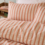 Hettie Printed Striped Duvet Cover Set in orange and pink, close-up of pillowcases and duvet with fine vertical stripes on a cream fabric ground, styled on a bed.