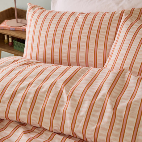 Hettie Printed Striped Duvet Cover Set in orange and pink, close-up of pillowcases and duvet with fine vertical stripes on a cream fabric ground, styled on a bed.