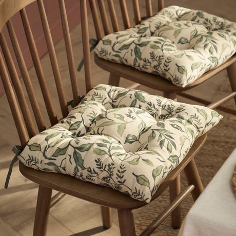 Herb Garden Linen Cotton Seat Pad in green, a tufted cream cushion with green leaf print and tie fastenings, shown on wooden spindle-back dining chairs.