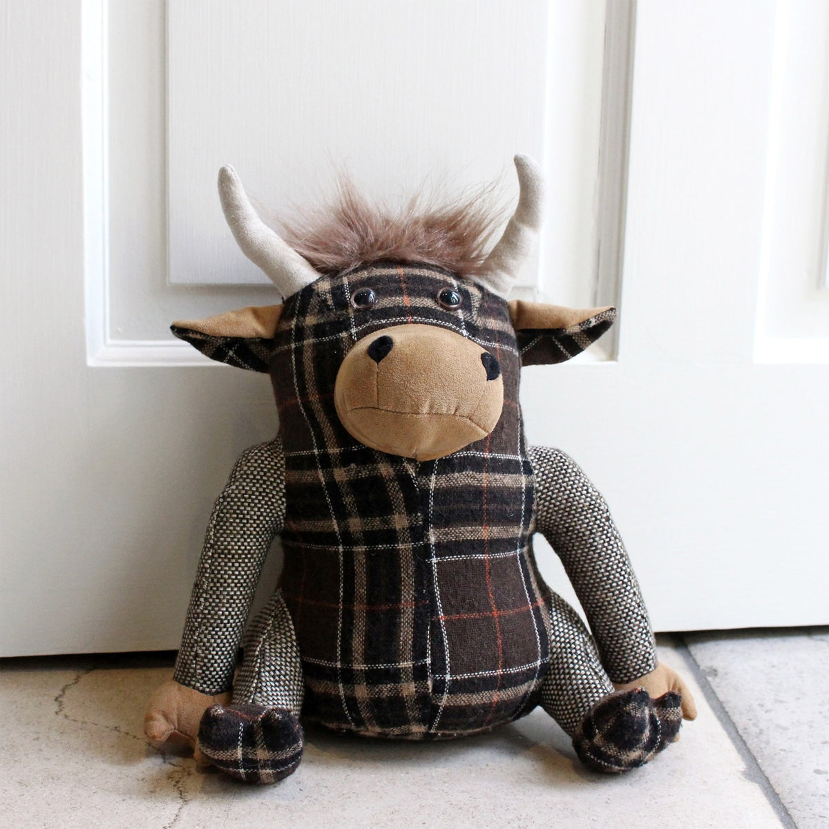Highland Cow Animal Polyester Door Stop in brown tartan fabric, seated figure with tan snout and hands, pale horns and tufted hair, shown against a white door.