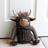 Highland Cow Animal Polyester Door Stop in brown tartan fabric, seated figure with tan snout and hands, pale horns and tufted hair, shown against a white door.