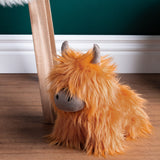 Highland Cow Velvet Faux Fur Door Stop in orange, shaggy fur with grey velvet snout and horns, shown on a wooden floor beside a chair leg.