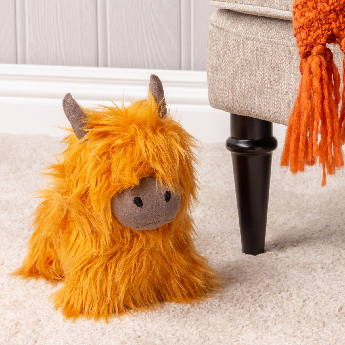 Highland Cow Velvet Faux Fur Door Stop in orange, shaggy fur with a grey velvet snout and horns, shown on carpet beside an upholstered armchair leg.
