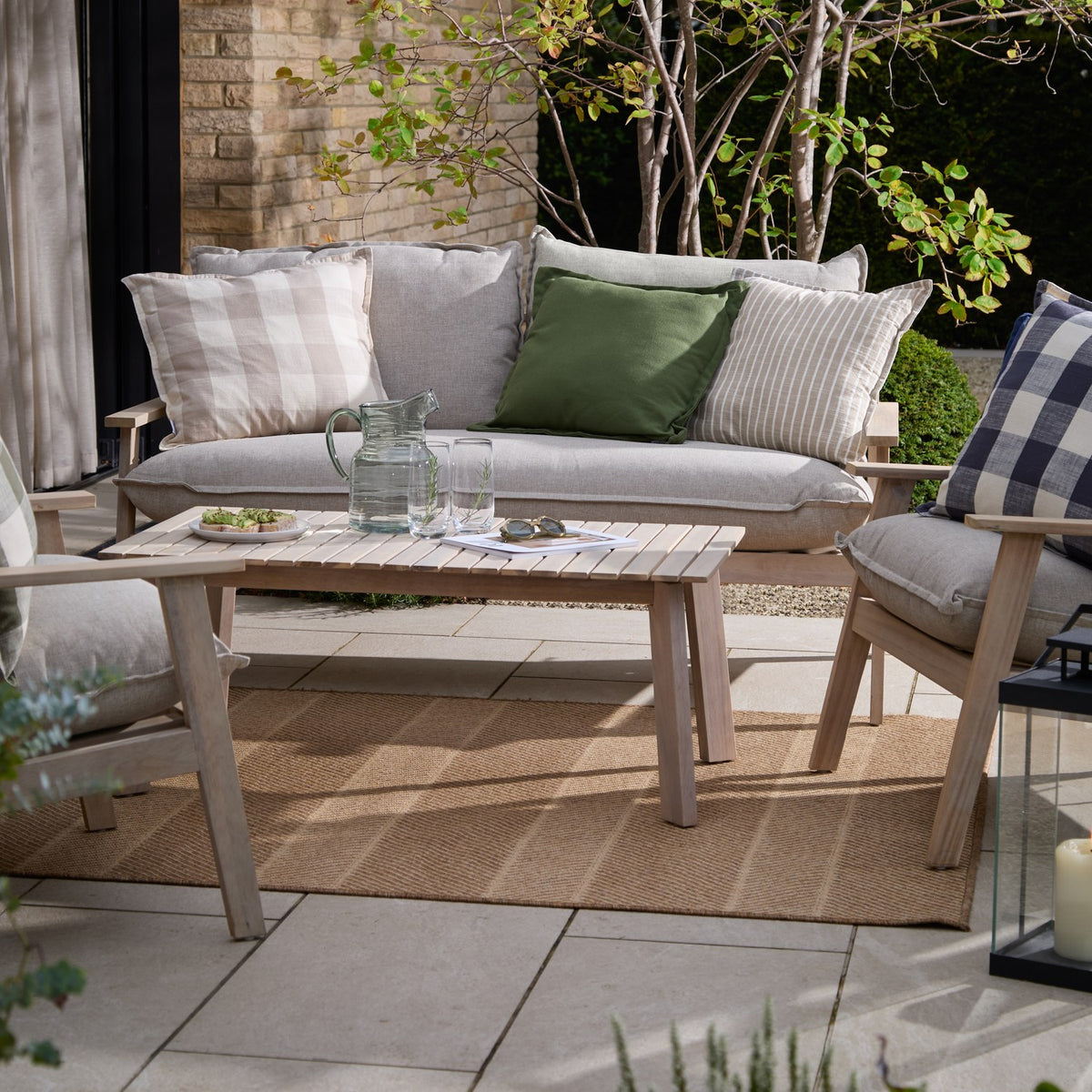 Holmfirth Canvas Plain Cotton Cushion in eucalyptus green on a grey outdoor sofa, styled with striped and check scatter cushions on a garden patio.