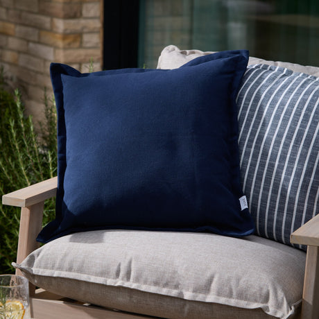 Holmfirth Canvas Plain Cotton Cushion in navy with a flanged edge, shown on an outdoor sofa with striped and linen-textured cushions in a garden setting.