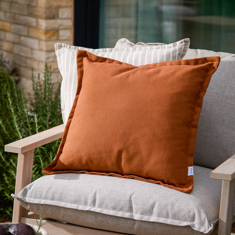 Holmfirth Canvas Plain Cotton Cushion in pecan orange, a square cushion with piped edges, shown on a light grey outdoor sofa alongside striped scatter cushions.
