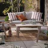 Holmfirth Canvas Plain Cotton Cushion in Pecan, a solid orange square cushion shown on a grey outdoor sofa alongside striped cushions on a patio setting.