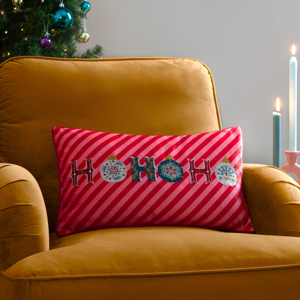 Rectangular velvet cushion with "HoHoHo" lettering and bauble motifs on a red and pink candy-striped ground, styled on a mustard armchair beside a Christmas tree.