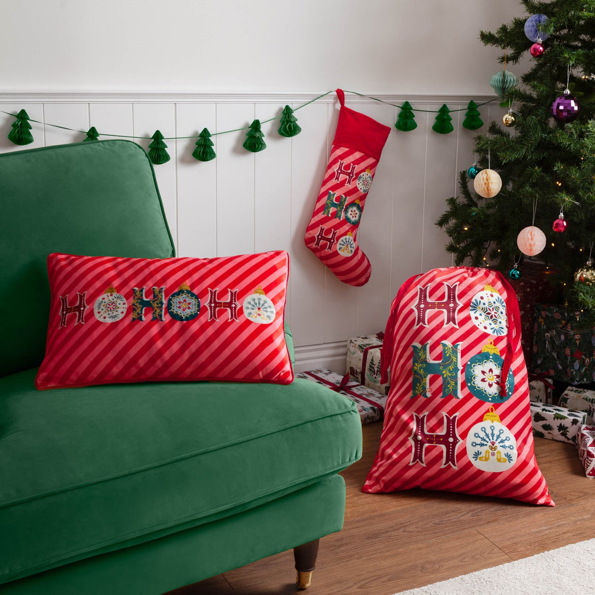 Rectangular red velvet cushion with candy-striped ground and "HoHoHo" slogan in ornate lettering with bauble motifs, shown on a green sofa beside a Christmas stocking and tree.